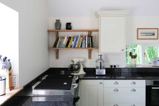 Bright kitchen with white cabinets, dark countertops, and a mixer. Shelves hold books and vases. A serene, tidy, and inviting cooking space.