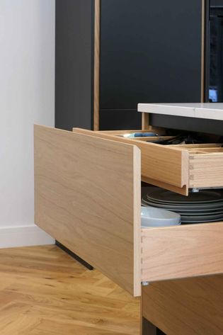 Open wooden kitchen drawers with plates and cutlery inside, set against a herringbone wooden floor and dark cabinetry.