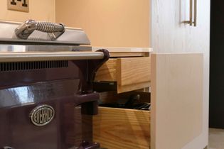 Purple stove beside a wooden kitchen drawer, slightly open, filled with utensils. The setting is warm and modern.
