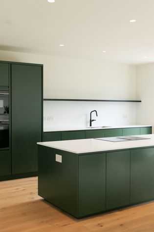 Modern kitchen with dark green cabinets, white countertops, and black tap. Wooden floor and recessed lighting create a modern, serene ambiance.