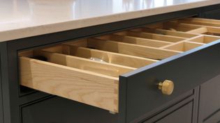 Open kitchen drawer with wooden cutlery organizer, featuring multiple compartments. The drawer has a dark exterior and a gold knob, set below a light countertop.