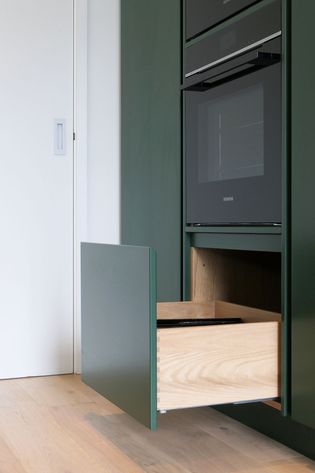 Modern kitchen with dark green cabinetry featuring an open wooden drawer beneath a built-in oven. Light wood flooring adds warmth.