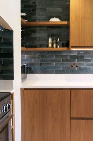 Modern kitchen with wood cabinets, white countertop, dark blue-grey tile backsplash, open shelves, and a partially visible oven.