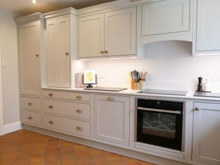 Shaker kitchen with pale grey cabinets, brass handles, and white countertops. Includes an oven, induction hob, utensils, and a decorative picture.