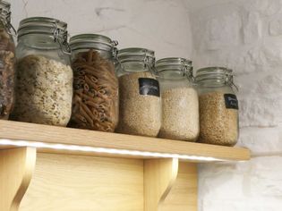 Glass jars filled with oats, pasta, quinoa, rice, and brown rice are neatly arranged on a wooden shelf against a white brick wall.