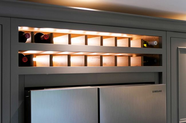 Illuminated wine rack above a stainless steel refrigerator, holding several wine bottles horizontally. Modern and sleek kitchen design.