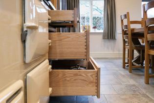 Open wooden kitchen drawers with pans inside, near a cream-colored stove. A window with curtains and a wooden dining table are to the side.