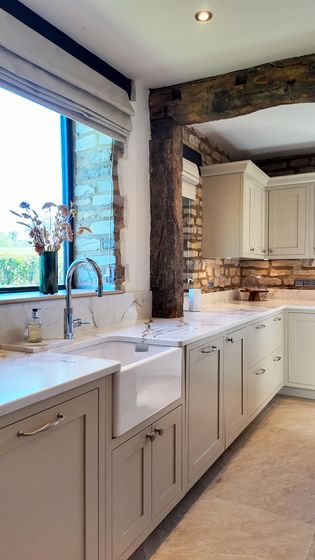 A Shaker kitchen featuring light-coloured cabinetry, a farmhouse sink, a stone wall, and a large window with natural light.