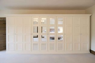 A large, elegant white-colored wardrobe with mirrored panels stands in a bright room. The reflection reveals a bed and chandelier, conveying a luxurious feel.