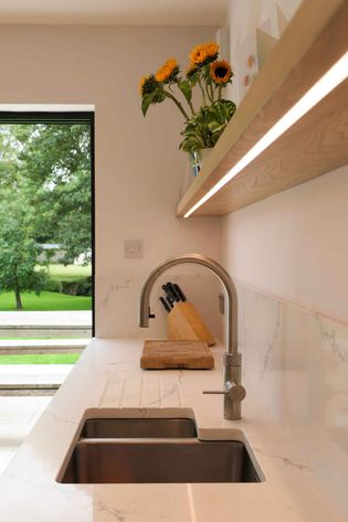 Modern kitchen with a white marble countertop, stainless steel sink, and sleek faucet. A knife block and sunflowers add warmth. Large window with garden view.