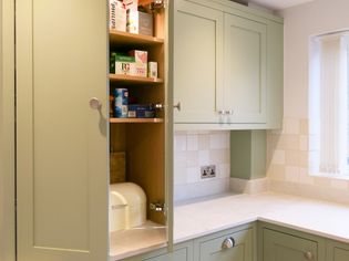 Open kitchen cabinet in a light green tone, displaying tea boxes and spices. Neutral countertop and cream tiles create a calm, organized ambiance.