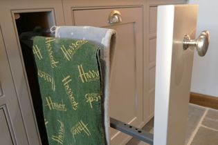 A pull-out kitchen cabinet displays a towel rack with a green towel labeled "Harrods." The cabinet is white with silver knobs, creating a modern look.