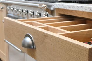 Open oak kitchen drawer with multiple slots next to a stainless steel oven. The drawer has a cup handle in iron material.