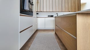 A modern kitchen with wooden accents, featuring a long rug on the floor, sleek cabinetry, and natural light filtering through wooden slats.