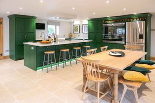 Spacious kitchen with green cabinets, white countertops, and stainless steel appliances. Central island with stools, dining table with colorful cushions.