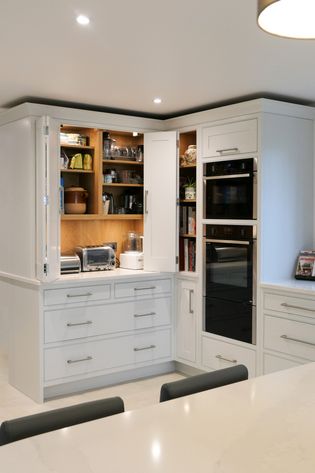 Shaker kitchen with white cabinetry, open pantry with organized shelves, built-in ovens, and marble countertops. Bright, functional, and tidy space.