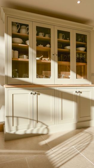 A light-coloured cabinet with glass doors displaying dish ware, set against a wall with shadows cast on the floor.