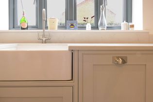 Bright kitchen with a large white farmhouse sink. Grey cabinets, a modern tap, and a window sill with decor, including a wine bottle and plant.