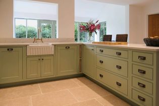 A cozy kitchen with sage green cabinets, a white farmhouse sink, and large windows. A vase of vibrant pink flowers adds a cheerful touch.