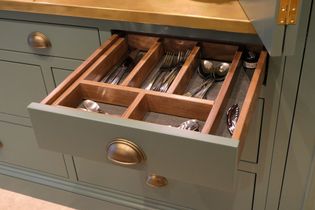 Open kitchen drawer with wooden dividers, neatly organized with silver cutlery including knives, forks, and spoons. Soft lighting, elegant feel.