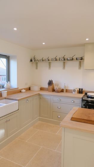 A Shaker kitchen with light-coloured cabinetry, a wooden countertop, and stainless steel appliances. Shelves display cookware, and there is a window providing natural light.
