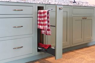 A light green kitchen cabinet with a pull-out towel rack, featuring red and white checkered towels. Granite worktop above. Clean and organized look.