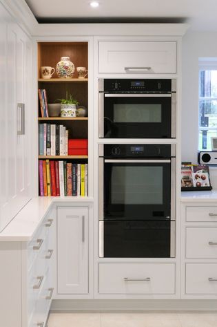 Shaker kitchen corner with white cabinets, a double oven, and a bookshelf filled with colorful cookbooks. A vase and mugs add warmth.