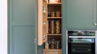 Open green pantry cabinet with jars of pasta, spices, and snacks on wooden shelves. Adjacent is a built-in oven. The scene is organized and tidy.