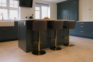 Shaker kitchen with a teal island and three grey bar stools with gold accents. Sunlight streams through windows, illuminating sleek cabinetry.
