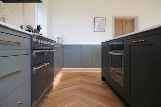 Shaker kitchen with sleek green and blue cabinets and brass handles. Features a stainless steel oven, wood flooring, wall art, and a warm, inviting ambiance.