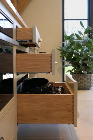 Wooden kitchen drawers are open, revealing pots and pans, against a backdrop of a large window and a potted plant.
