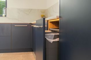 Modern kitchen with dark blue cabinets, sleek handles, and under-cabinet lighting. A partly open door reveals an integrated bin.