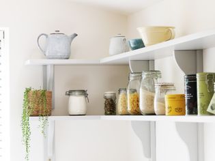 Open white shelves displaying a teapot, jars of dry goods, and a trailing plant. The arrangement is organised clean.