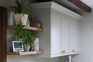 A kitchen corner featuring neutral cabinets with gold handles and two wooden shelves. The shelves hold plants, a digital clock, and storage items.