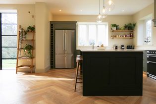 Shaker  kitchen with light wood flooring, a dark island, and pendant lights. Lush plants on shelves create a fresh, inviting feel with natural light coming in.