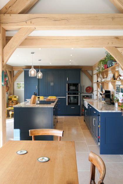 Rustic Shaker kitchen with exposed wood beams, navy cabinetry, granite countertops, pendant lights, open shelves, and a wooden dining table.