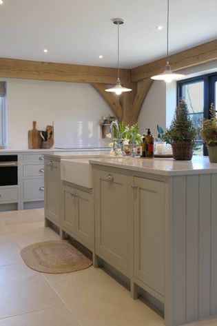 Farmhouse kitchen with cream shaker cabinets, a large farmhouse sink, and wooden ceiling beams. Bright lighting and potted plants add warmth.