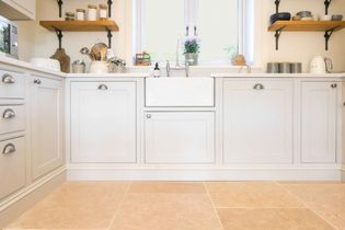 A shaker kitchen with light grey cabinets, a farmhouse sink, and a window above. Shelves hold jars and cups, creating a warm, inviting atmosphere.