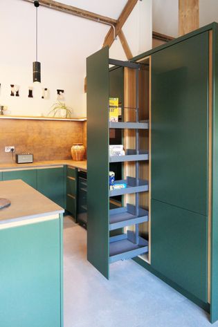 A modern kitchen featuring green cabinetry, open space tower with pantry items inside and warm lighting.