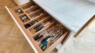 A wooden kitchen drawer filled with various utensils, including knives, spoons, scissors, a measuring spoon, a strainer, and other tools, all neatly organised within compartments.