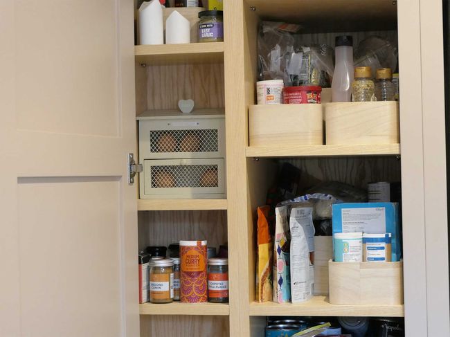 A wooden pantry cupboard with open doors shows neatly organized shelves. Various spices, jars, and packaged goods are stored in baskets, giving a tidy appearance.