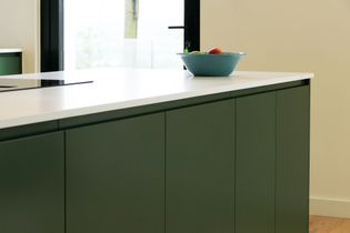 Modern kitchen with sleek green cabinets under a white countertop. A blue bowl with fruit is on the counter, near a glass door. Bright and clean ambiance.