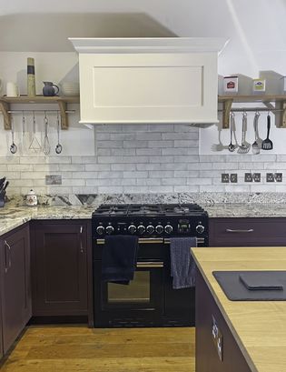 A Shaker kitchen featuring a black stove with multiple burners, a white range hood above, granite countertops, and wooden shelves with kitchen utensils.