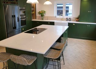 Stylish kitchen with green cabinets, white countertops, and wicker bar stools. Bright lighting, flowers, and decor create a cozy, modern vibe.