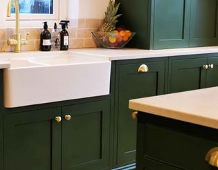 Elegant kitchen featuring dark green cabinets with gold knobs, a white farmhouse sink, gold faucet, and a fruit bowl on the countertop.