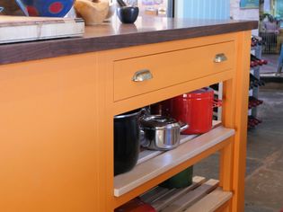 Bright orange kitchen island with wooden top, highlighting a drawer and lower open shelf holding colorful pots. Warm and inviting atmosphere.