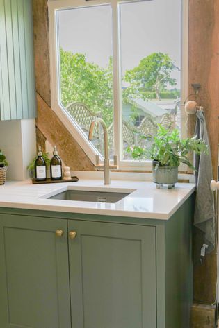 A kitchen sink with a sleek faucet is situated under a large window, surrounded by greenery. Green cabinets and countertop bottles create a fresh, serene vibe.