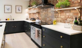 A shaker kitchen with dark grey cabinets, a brick backsplash, and open wooden shelves displaying plants. A stove with a kettle adds a cozy touch.