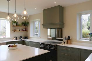 Shaker kitchen with green cabinets, white worktops, and a central island. Bright pendant lights hang above. Windows and open shelves add brightness.
