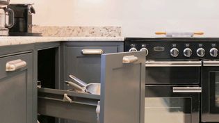 Dark grey shaker kitchen cabinets are open, with cooking utensils visible in a drawer. A black oven with silver knobs stands nearby.